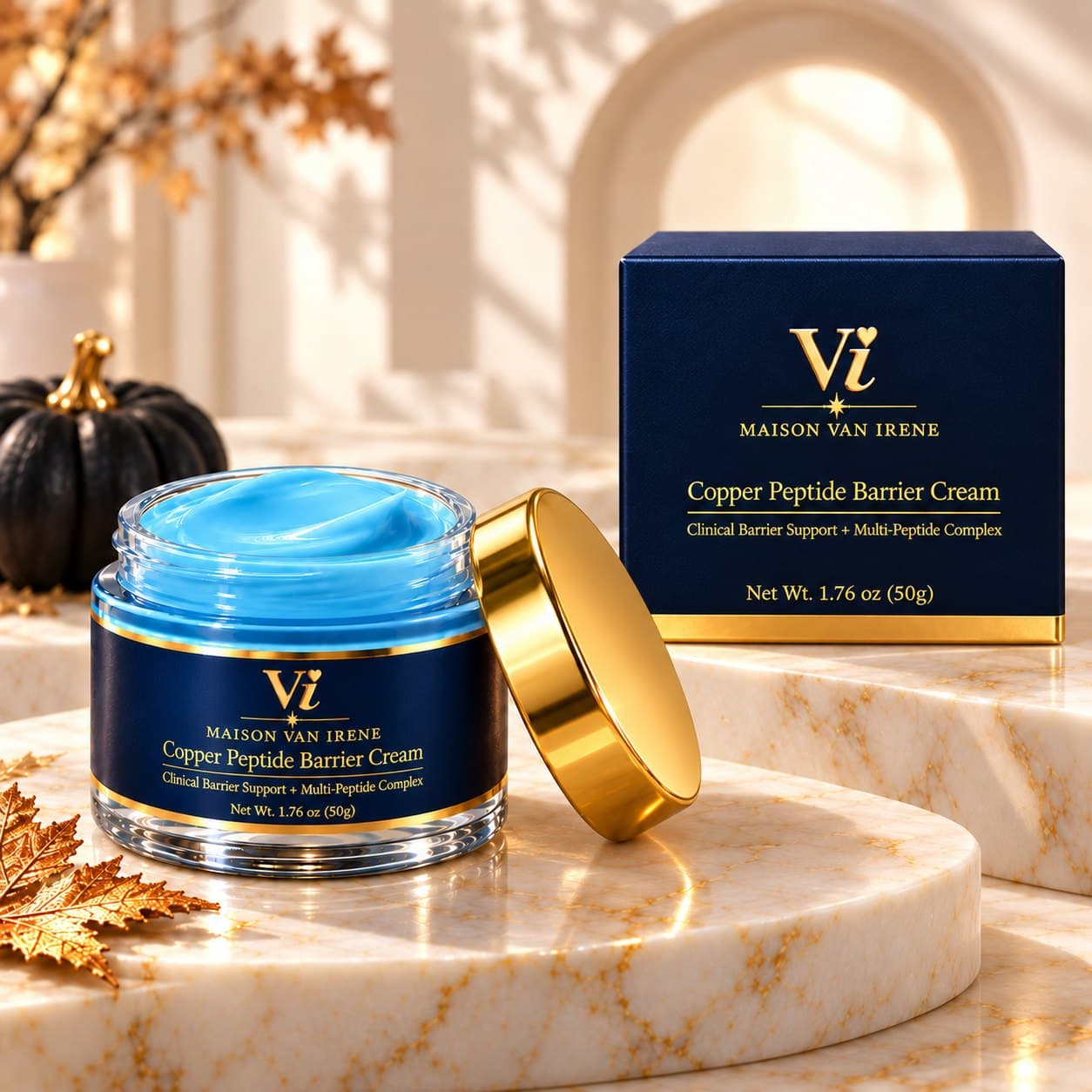 Maison Van Irene Copper Peptide Barrier Cream open jar with polished gold lid and navy carton, gold autumn leaves and a white pumpkin on marble